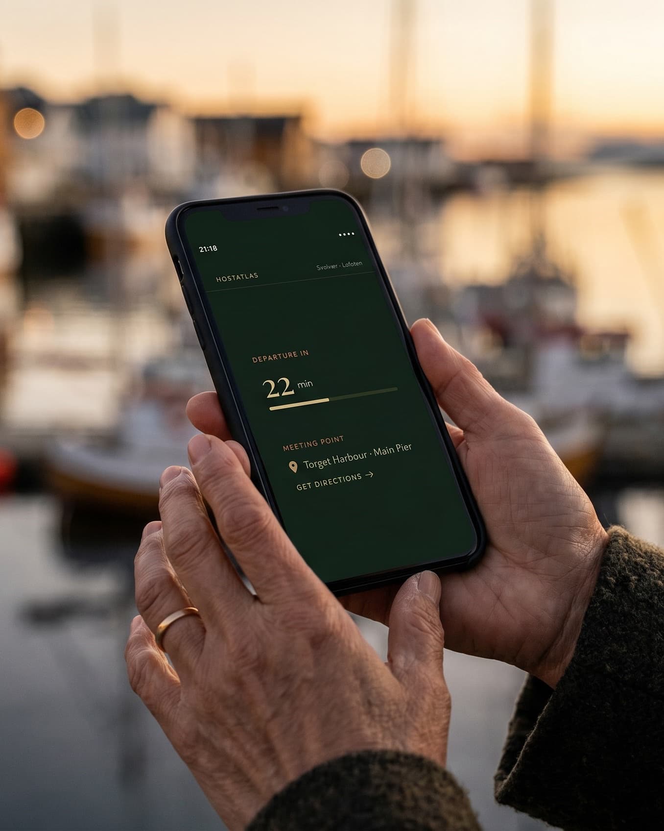 A guest's hands holding a phone showing the HostAtlas departure countdown, golden-hour harbour behind
